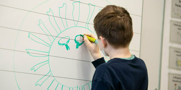 Ein Kind steht an einer Tafel mit dem Rücken zur Kamera. Mit einem Stift malt es einen großen Kreis mit vielen Zacken, die Umrisse des Coronavirus