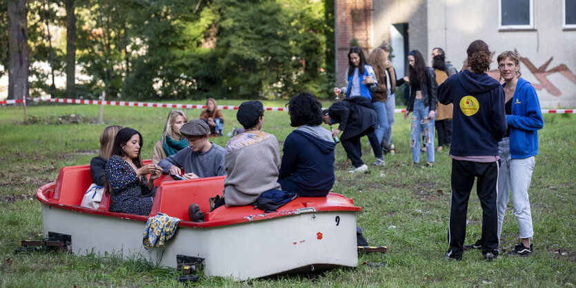 Ein Boot liegt auf einer Wiese und mehrere Menschen sitzen entspannt drauf und stehen drum herum.