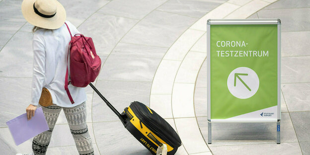 Eine Person zieht einen Rollkoffer hinter sich her, neben ihr ein Schild, auf dem "Corona Test-Zentrum" steht.