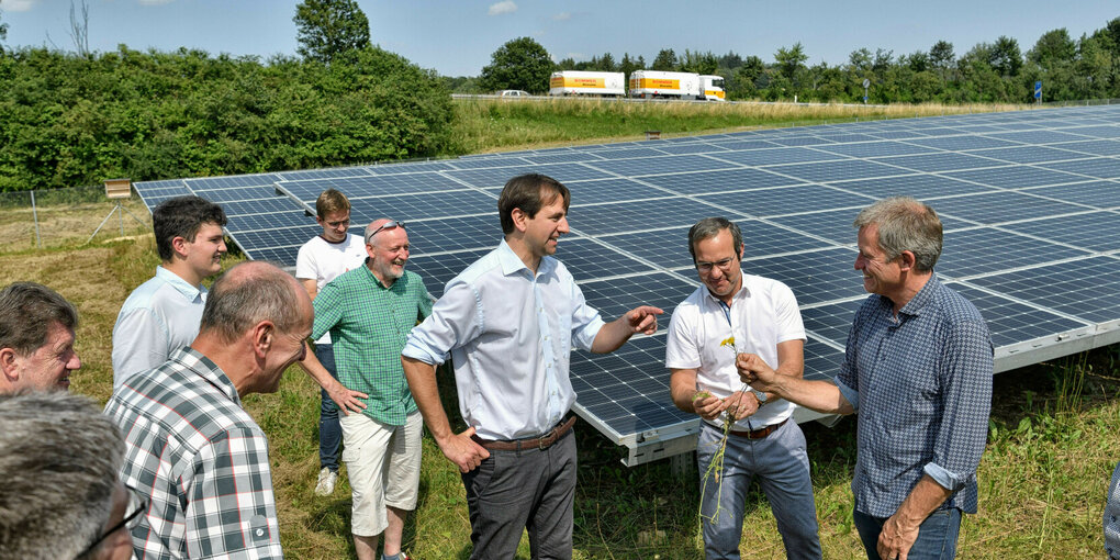 Eine Gruppe von Männern steht vor Solaranlagen auf einer Wiese, direkt dahinter liegt die Autobahn