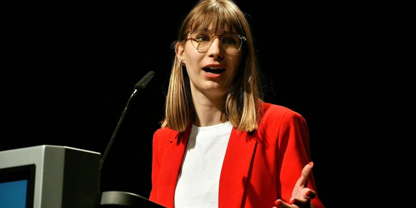 Jeanne Dillschneider steht an einem Rednerpult und trägt ein rotes Sacko