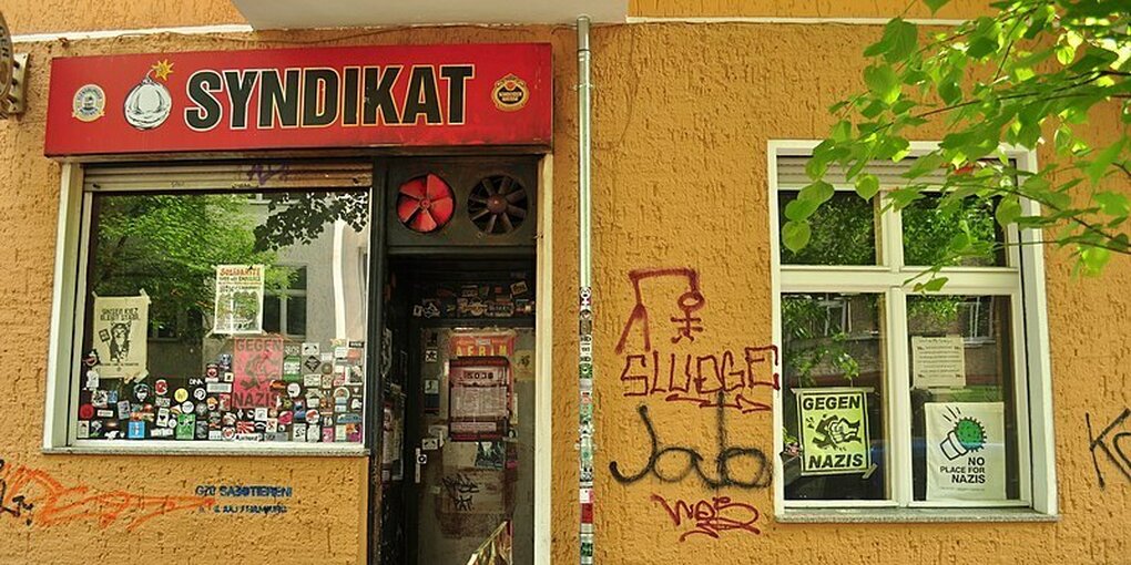 Eine gelbe Hauswand, darin ein Schaufenster und eine Kneipentür. Darüber das rote Schild 'Syndikat'