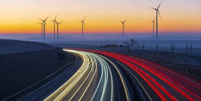 Das Bild zeigt eine Autobahn und im Hintergrund einen Windpark im Sonnenuntergang.