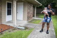 Eine Frau mit Baby auf dem Arm steht vor einem Haus.