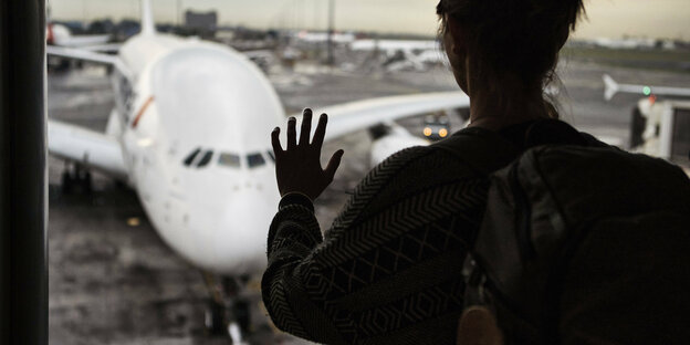 Eine Hand an einem Fenster, im Hintergrund ein Flugzeug.