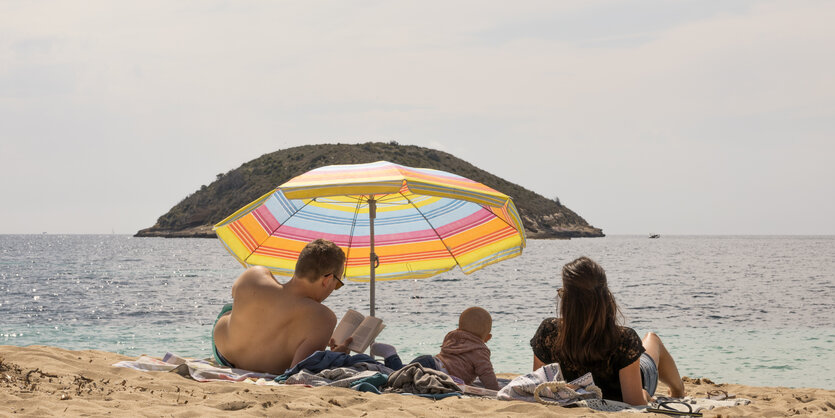 Eine Familie liegt am Strand unter dem Sonnenschirm und schaut aufs Meer. Das Baby liegt in der Mitte