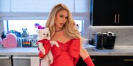 Paris Hilton steht in einem roten Kleid mit einem weißen Hund im Arm in einer Küche