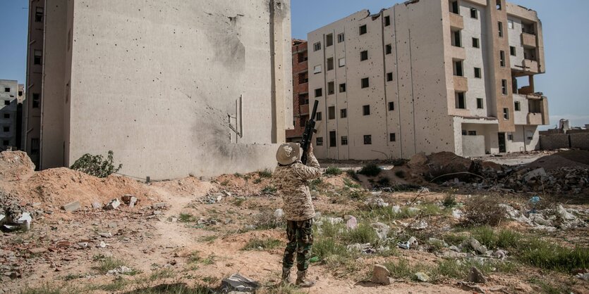 Ein Mann, vor Hausruinen stehend, schießt mit einem Gewehr in die Luft