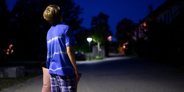 Ein kleiner Junge mit einem blauen T-Shirt und einem roten Kissen unter dem Arm, läuft in der Nacht über eine Straße