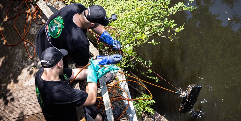 Zwei Männer mit Basecaps und Handschuhen ziehen etwas von einer Brücke aus dem Wasser