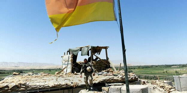 Ein Bundeswehrsoldat auf einer Barriere aus Sandsäcken in Afghanistan. Im Vordergrund weht eine deutsche Fahne