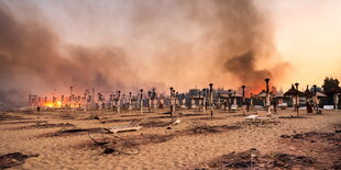 Strandansicht mit verkohlten Sonnenschirmen und Liegen, im Hintergrund offenes Feuer