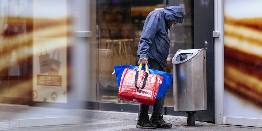 Ein Mann schaut in einen Mülleimer, er hat zwei Plastiktaschen in der Hand