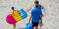 Vater, Mutter Kind gehen mit buner Luftmatraze am Strand entlang