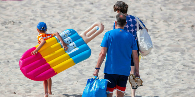 Vater, Mutter Kind gehen mit buner Luftmatraze am Strand entlang
