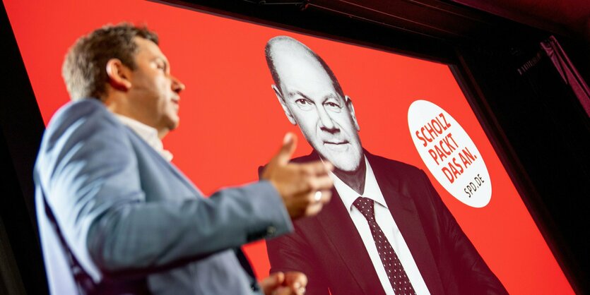 Ein Mann im Anzug vor einem großen roten SPD-Plakat mit einem grimmig schauenden Mann darauf