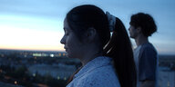 Zwei junge Mneschen stehen auf einem Hausdach und blicken im Abendhimmel über eine Großstadt