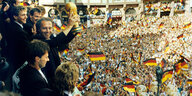 die deutsche Fußball-Nationalmannschaft der Männer von 1990 feiert ihren WM-Sieg, Franz-Beckenbauer hält den Pokal über einer Ansammlung von feiernden Menschen und einem Meer an Deutschlandfahnen in die Luft,