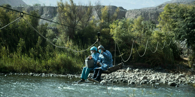 Zwei mit medizinscher Schutzkledung ausgestattete Personen werden an einem Seil über einen Fluss gezogen