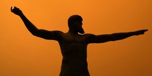 Die Statue von Posseidon mit ausgestreckten Armen vor einem glutroten Hintergrund
