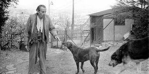 Ein schwarzweiß--Foto, man sieht einen dürren Mann, Céline, in Kleidern, die ihm zu groß sind, mit zwei Hunden