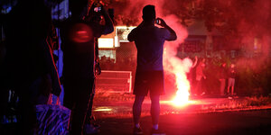 Zwei Männer filmen mit ihren Handys ein Feuer, dahinter Demonstranten