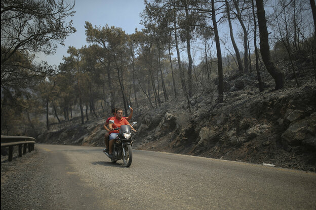 Zwei Männer fahren auf einem Motorrad durch eine verbrannte Landschaft, Der Mann auf dem Sozius fotografiert mit seinem Handy