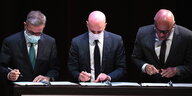 Drei Männer beugen sich nebeneinander über Dokumente, die zur Unterschrift vorliegen