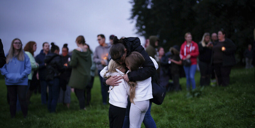 EIn Park. Eine Frau umarmt zwei kleine Mädchen. Hinter ihr sind weitere Menschen versammelt.