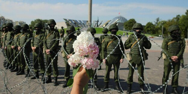 Eine Hand hält eine Blume hoch vor einer Reihe von Polizisten hinter Stacheldraht