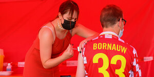 Ein Mann in einem roten Trikot (Aufschrift "Bornauw – 33") bekommt von einer Frau in rotem Kleid eine Impfung in den Oberarm. der hintergrund ist auch rot.