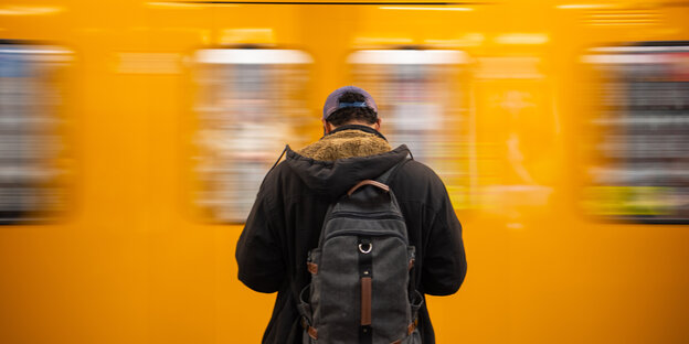 Ein Mann ist von hinten zu sehen wie er auf eine Berliner U-Bahn wartet, die gerade einfährt.