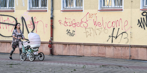 Eine Frau mit Kinderwagen läuft vor einer verschmierten Wand "Refugees Welcome" mit einem NOT dazwischen