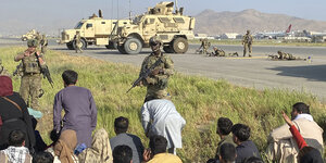 US-Soldaten bewachen eine Absperrung am internationalen Flughafen. Man sieht mehrere bewaffnete Soldaten auf einem Grasstreifen, hinter ihnen Militarfahrzeuge und das Rollfeld. Vor ihnen kauern Afghanen im Gras