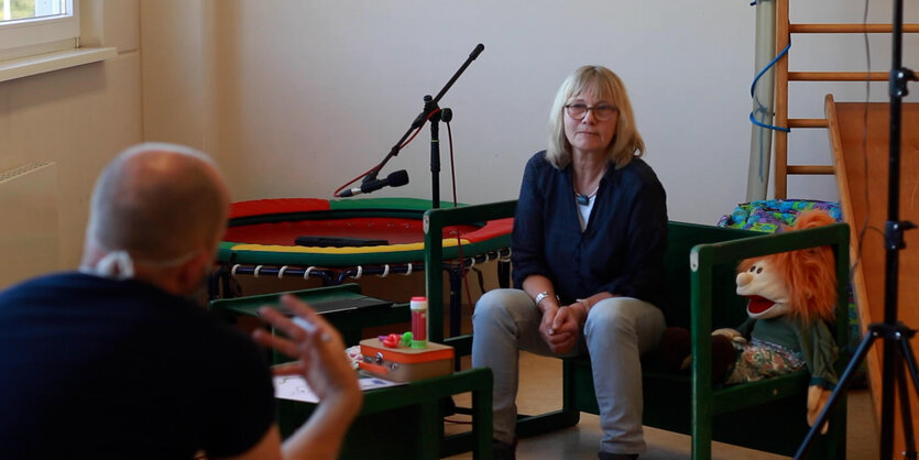 Eine Frau sitzt in einem Zimmer mit Spielsachen und wir von einem Mann, links im Bild, interviewt.