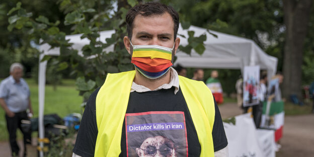 Ein Teilnehmer bei einer Demonstration gegen den Iran mit gelber Warnweste, Regenbogen-Mundschutz und "Dictator kills Iran"-T-Shirt