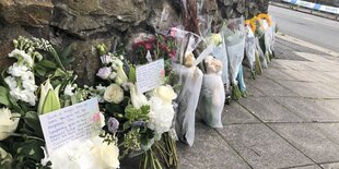 Blumen lehnen an einer Steinmauer. Zwischen den Blüten sind Briefe. Auf dem ersten Brief erkennt man die Schrift: "Such a tragic loss of lives. You will not be forgotten. Our deepest sympathy goes to the families and loved ones at this sad time."