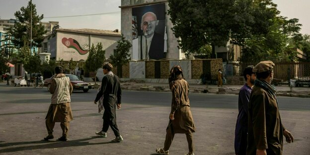 Männer gehen eine Straße entlang, im Hintergrund ist eine Gebäude an dem ein großes Plakat mit dem Portrait von Ashraf Ghani hängt