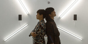 Das Duo Nguyễn + Transitory steht Rücken and Rücken vor eine Wand mit vier Lichtröhren