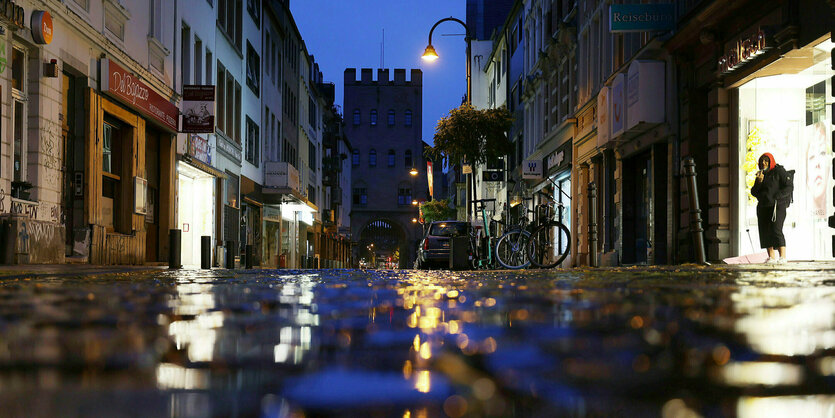 Eine enge Straße in der Kölner Innenstadt, auf einer Pfütze auf dem Kopfsteinpflaster spiegelt sich der Himmel