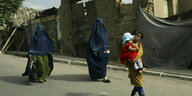 Zwei komplett verschleierte Frauen und ein unverschleiertes Mädchen mit einem Baby auf dem Arm laufen die Straße herunter, im Hintergrund eine Ruine