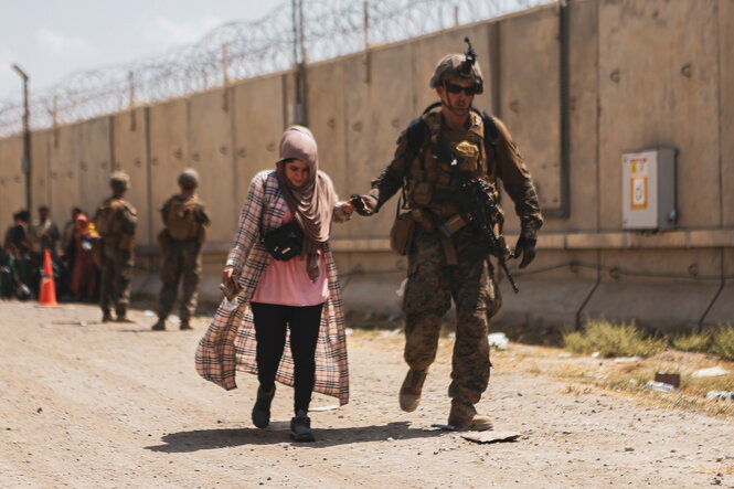 Ein US-Soldat hält eine afghanische Frau an der Hand. Im Hintergrund eine Mauer mit Stacheldraht.