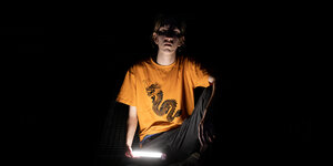 Ein junger Mensch mit einem orangenen T-Shirt, auf dem ein Drache abgebildet ist, sitzt im Dunkeln, nur angestrahlt durch einen Leuchtröhre, die er in der Hand hält