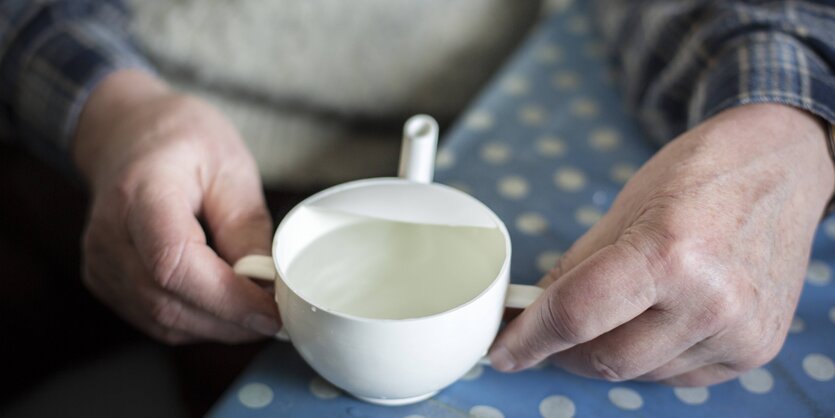 Hände eines alten Mannes halten eine Schnabeltasse, die auf eien gepunkteten Tischdecke steht