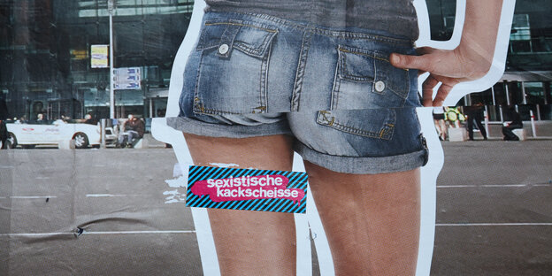 Ein Werbeplakat mit einer Frau in einer kurzen Shorts von hinten, über dem ein Sticker mit den Worten "Sexistische Kackscheiße" klebt.
