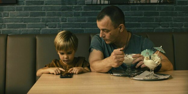 Milan (Ben Winkler, l.) und sein Vater Mario (Paul Wollin, r.) sitzen an einem Tisch. Vor Milan liegt ein Handy, vor Mario stehen zwei Becher Eis.