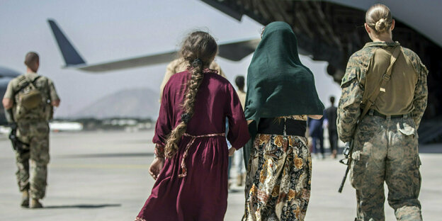eine Soldatin in Uniform bringt zwei Frauen oder Mädchen zum Flughafen. Die beiden anderen Personen tragen Kleider.