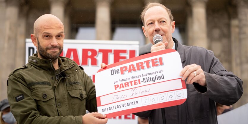 Marco Bülow und Martin Sonneborn stehen vor dem Bundestag und halten gemeinsam einen großen Mitgliedsausweis Marco Bülows mit der Nummer 50.000.