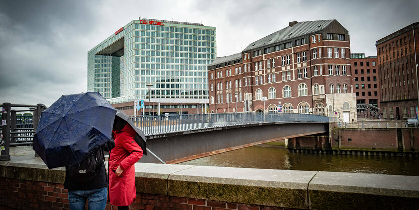 Zwei Menschen stehen am Wegesrand, man erkennt ihre Gesichter nicht, weil Regenschirme ihre Gesichter verdecken. Im Hintergrund ist das Verlagshaus des "Spiegel" zu erkennen