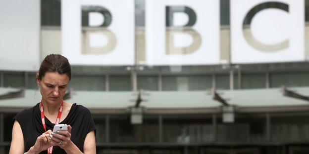 Frau steht vor dem BBC-Gebäude und schaut auf ihr Handy